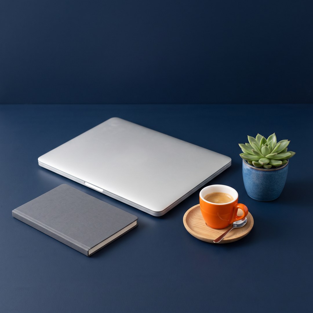 A closed laptop, a grey notebook, an orange espresso cup on a wooden saucer, and a small succulent on a dark blue desk.